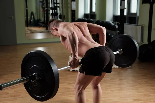 A muscular guy in sports shorts and a naked torso stands in the gym lifting a heavy barbell. — Stock Photo, Image