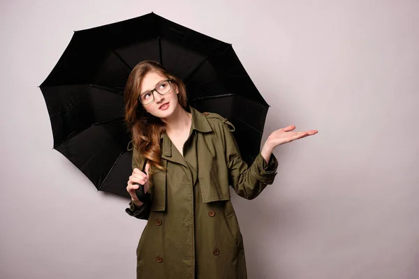 Uma menina com um manto verde e óculos fica com um guarda-chuva preto e, abrindo a mão, olha para o topo, inclinando a cabeça para o lado . — Fotografia de Stock