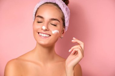 Kapalı gözlü, saçları toplanmış, yüzünde kremayla pembe bir arka planda duran bir kızın yakın portresi..