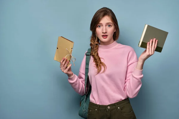 Egy lány egy pulóverben egy hátizsákkal áll, és meglepetten néz a kamerába, könyveket tart a kezében.. — Stock Fotó