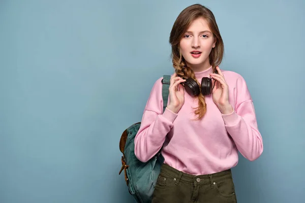A girl in a pink sweater with a backpack stands on a blue background and holds headphones with hands on her neck. — Stockfoto