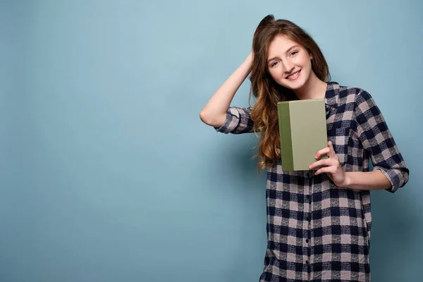 Ein junges Mädchen im karierten Kleid steht vor blauem Hintergrund und hält ein Buch vor sich, während sie ihre Hand durch ihr Haar streicht. — Stockfoto