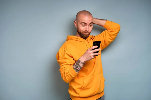 A bearded guy in a yellow sweatshirt, opening his eyes wide and pursing his lips, looks into a smartphone scratching his head
