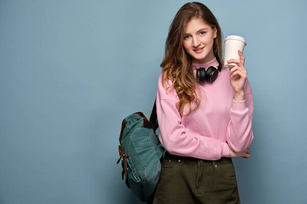 A young girl in a pink sweater and headphones with a backpack stands on a blue background with a plastic cup in her hand.