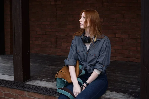Vörös hajú lány ül egy fa platformon vállpánttal és egy hátizsák, úgy néz ki, az oldalán — Stock Fotó