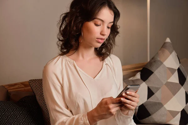 Portrait of a dark-haired curly girl in a light blouse sitting among pillows and looking at the phone.