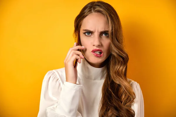 A girl with curls and red lipstick in a white blouse stands on a yellow background and grimacing angrily speaks on the phone. — Stockfoto