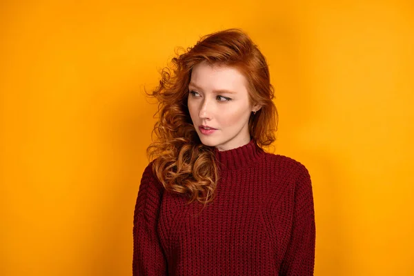 Egy vörös hajú lány burgundi pulóverben áll sárga háttérben, és oldalra néz.. — Stock Fotó