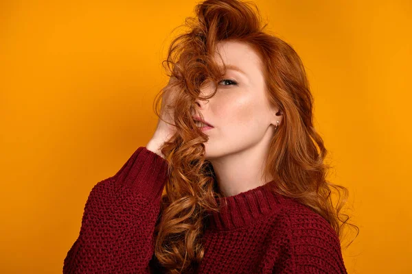 Mooi roodharig meisje in een bordeaux trui kijkt naar de camera die haar gezicht bedekt met gedweild haar op een gele achtergrond — Stockfoto