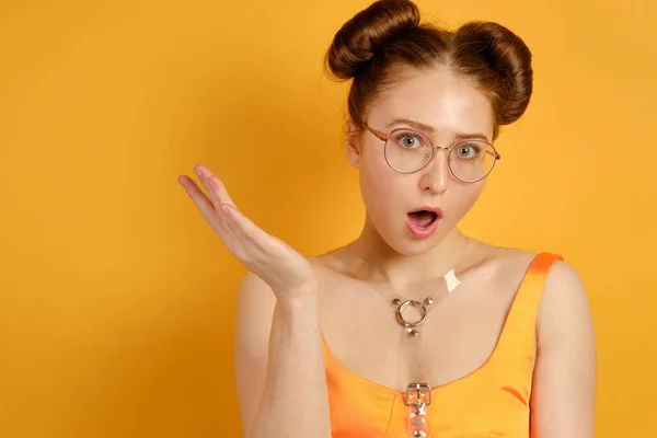 Uma menina em um top laranja e óculos redondos fica em um fundo amarelo, olha para a câmera, abrindo a boca e levantando a mão — Fotografia de Stock