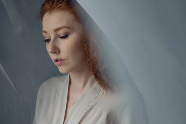 Uma menina ruiva fica meia volta entre as cortinas e olha para baixo . — Fotografia de Stock