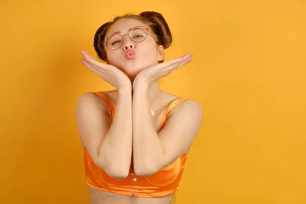 En rödhårig flicka i orange topp och runda glasögon står på en gul bakgrund, skickar en kyss, sätter hakan på öppna handflator — Stockfoto