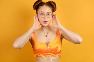 Turuncu bluzlu ve yuvarlak gözlüklü kızıl saçlı bir kız kameraya bakıyor, yüzünün yanında uzanmış avuç içlerini tutuyor.