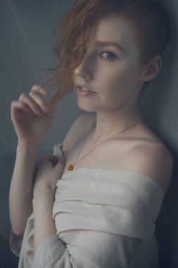 Çıplak omuzlu kızıl saçlı bir kız kameraya bakar, yüzünün bir kısmını saçlarla kaplar. Perdenin arkasını çevirin..