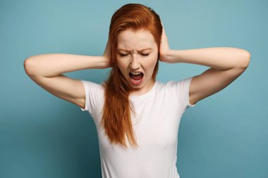 Kızıl saçlı bir kız mavi bir arka planda beyaz bir tişörtle duruyor, kulaklarını elleriyle sıkıyor ve gözlerini kısarak haykırıyor.