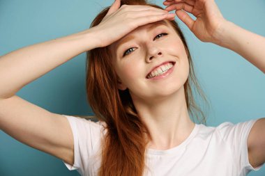 Kızıl saçlı kız mavi bir arka planda beyaz bir tişörtle duruyor, gülümsüyor, alnında bir avuç içi ile..