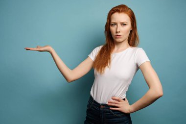 Kızıl saçlı kız beyaz tişörtlü mavi bir arka planda duruyor, avuç içi ve çerçeve kaş çatık.