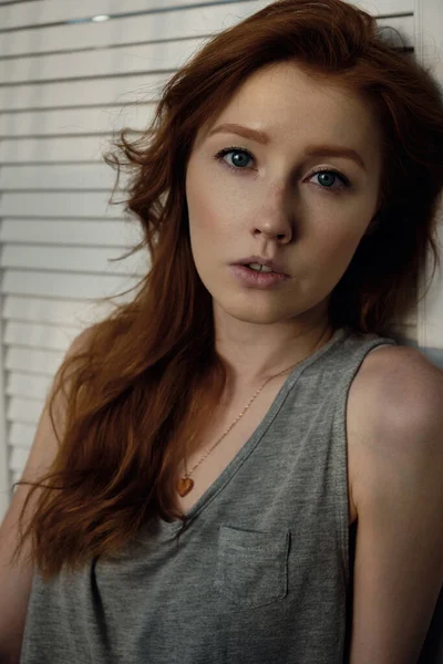Μια όμορφη κοκκινομάλλα με τα μαλλιά της και φακίδες σε ένα γκρι μπλουζάκι κοιτάζει την κάμερα, ακουμπισμένη σε μια λευκή ξύλινη οθόνη — Φωτογραφία Αρχείου