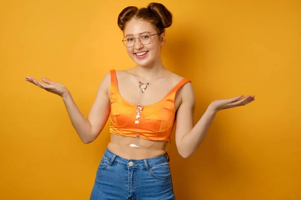 Das Mädchen mit zwei Strahlen, Brille und orangefarbenem Oberteil steht auf gelbem Grund und lächelt und breitet die Handflächen seitlich aus lizenzfreie Stockfotos