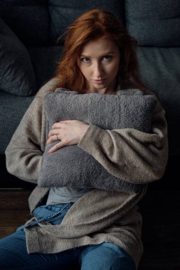 Sıcak kazaklı kızıl saçlı bir kız oturuyor, gri bir yastığa sarılıyor ve yukarıdan kameraya bakıyor.