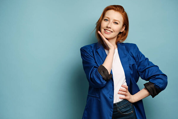 Red-haired girl in a blue jacket stands on a blue background and smiles, raising his hand to face.