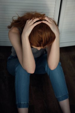 Kırmızı saçlı, tişörtlü ve pantolonlu bir kız kafası aşağıda, parmakları saçında, yukarıdan çerçeveli bir şekilde yerde oturuyor..