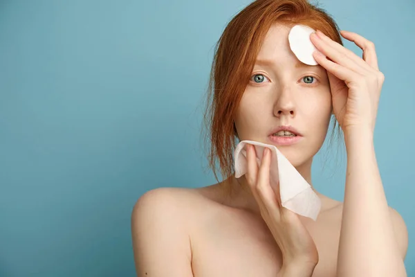 Redhead girl with clean radiant skin, without makeup, wipes her face with a napkin and cotton pad, standing on a blue background