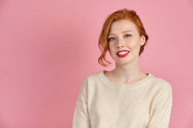 Açık renk süveter giyen, kırmızı dudaklı güzel kızıl saçlı kız pembe arka planda duruyor ve geniş bir şekilde gülümsüyor.