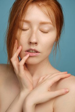 Kızıl saçlı, parlak tenli, makyajsız, gözleri kapalı, mavi arka planda duran, parmaklarını yüzüne koyan bir kız.