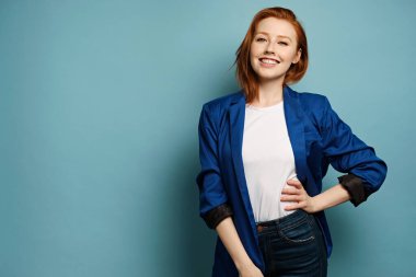 Beyaz tişörtlü, mavi ceketli, kızıl saçlı bir kız mavi arka planda duruyor ve geniş bir gülümsemeyle elini beline koyuyor.