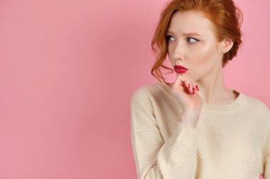 Açık renk kazaklı, pembe dudaklı, kızıl saçlı bir kız pembe bir arka planda duruyor ve yanlara bakıyor, çenesini destekliyor..