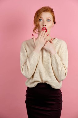 Kırmızı dudaklı kızıl saçlı bir kız, pembe arka planda duruyor, kırmızı tırnaklı bir avuç içi ile şaşırtıcı bir şekilde açık bir ağız kapatıyor.