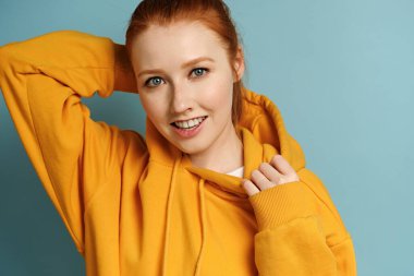 Sarı tişörtlü kızıl saçlı bir kız mavi arka planda duruyor ve neşeli bir şekilde çerçeveye bakıyor, kaputu ayarlıyor.