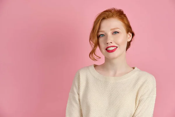 Gyönyörű vörös hajú lány, világos pulóverben, piros ajkakkal, rózsaszín háttérrel és széles mosollyal. — Stock Fotó