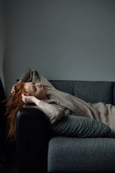 Bej rengi hırkalı kızıl saçlı bir kız kanepeye uzanmış, başını koltuğa yaslıyor.. — Stok fotoğraf