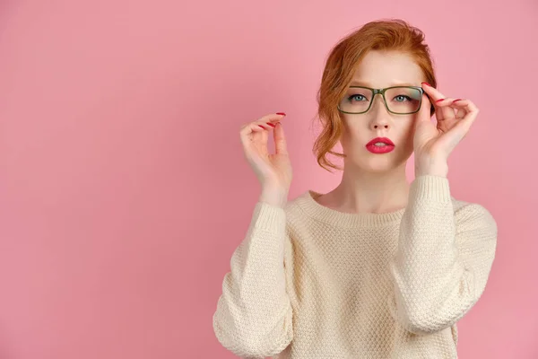 Schöne rothaarige Mädchen in einem hellen Pullover und mit roten Lippen steht auf einem rosa Hintergrund und setzt eine Brille auf. — Stockfoto