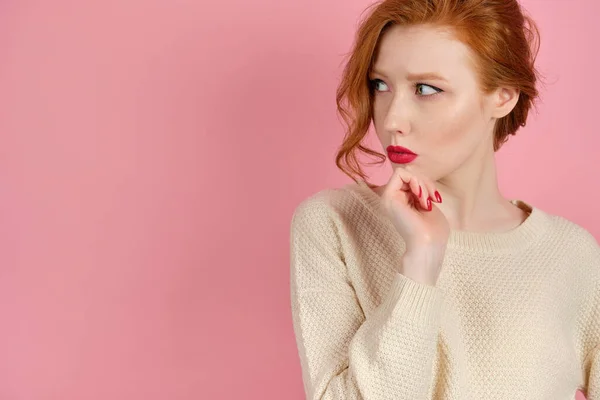 Egy vörös hajú lány, világos pulóverben, piros ajkakkal, rózsaszín háttérben áll, és óvatosan néz oldalra, az állát támasztva.. — Stock Fotó