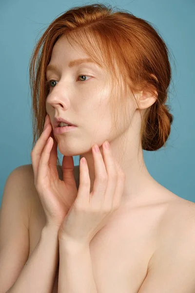 Chica pelirroja con la piel radiante limpia, sin maquillaje, de pie en una media vuelta sobre un fondo azul, poniendo sus palmas a la cara — Foto de Stock