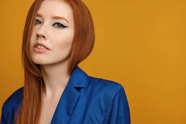 Portré egy vörös hajú lány hosszú nyilakkal és egy kék kabát áll egy félfordulat egy sárga háttér. — Stock Fotó