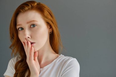 Temiz tenli, mavi gözlü, beyaz tişörtlü güzel kızıl saçlı kız çerçevenin içine bakar, dudaklarını parmaklarıyla kapatır.