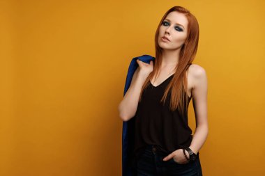  Siyah bluzlu ve mavi smokinli bir kız sarı arka planda duruyor, omzundan bir ceket atıyor ve elini cebine sokuyor.