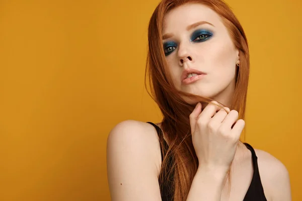 Chica pelirroja en un top negro y con maquillaje de ojos azules se levanta sobre un fondo amarillo y mira en el marco, jugando con el pelo — Foto de Stock