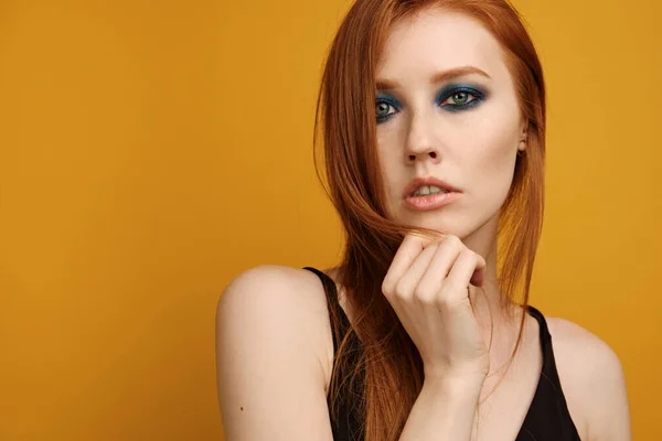 Hoofdschot. Het roodharige meisje in een zwart topje en met blauwe oogmake-up staat op een gele achtergrond en kijkt in het frame — Stockfoto