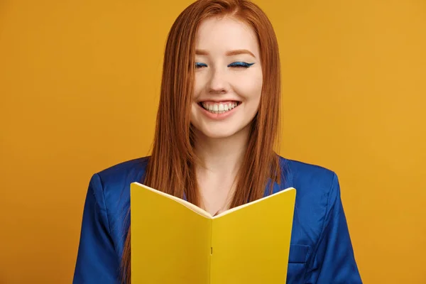파란색 재킷을 입고 밝은 화살표를 한 빨간 머리 소녀가 노란색 배경에 서서 노트북을 들고 활짝 미소를 짓고 눈을 감고 있습니다 — 스톡 사진