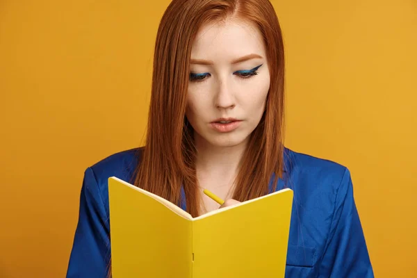 Piros hajú lány kék kabátban, fényes nyilakkal áll a sárga háttér és ír egy sárga notebook — Stock Fotó