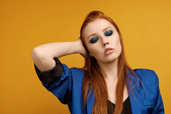 Roodharig meisje in een jasje en met blauwe ogen make-up staat op een gele achtergrond, stromende hand door haar en naar beneden kijkend — Stockfoto
