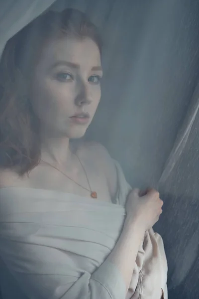 Piros hajú, kék szemű lány áll egy fehér áttetsző függöny előtt, és belenéz a keretbe, a függönyön keresztül lát. — Stock Fotó