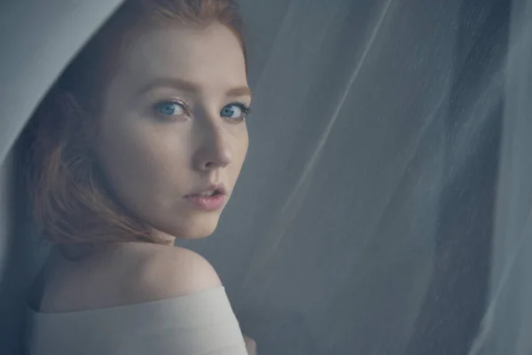 Uma menina de cabelos vermelhos com olhos azuis fica no meio de uma cortina translúcida e olha por cima do ombro, uma vista através da cortina . — Fotografia de Stock