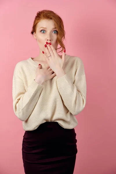 Vörös hajú lány, fehér pulóverben, rózsaszín háttéren áll, eltakarja a száját, és a szívéhez szorítja a kezét. — Stock Fotó