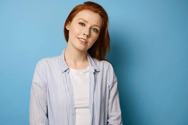 Red-haired girl in a striped shirt and a white T-shirt stands on a blue background with a slight smile and bowing head to the side